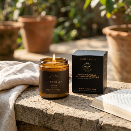 A lit amber glass candle jar labeled Citronella by Lumère Roots sits on a stone surface beside its black product box, with a folded cream cloth and an open book in a sunlit outdoor garden setting.