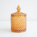An amber-colored glass jar with a matching lid, featuring a textured diamond-cut pattern and a small finial on top, set against a plain white background.